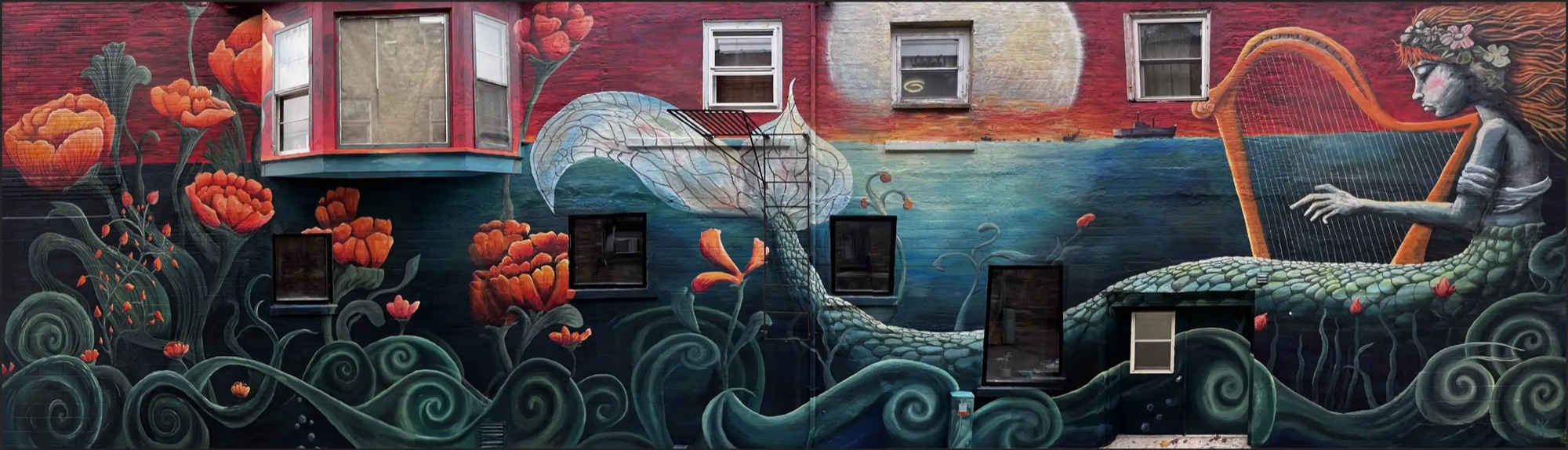 Harbor mermaid mural, Algoma: a full building face painted around its windows — a mermaid playing a golden harp at moonrise, her scaled tail winding through waves and orange poppies.